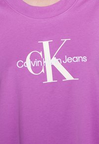 Camiseta de algodón morada con el texto "Calvin Klein Jeans" en blanco y un gran logo "CK" en una fuente moderna. Diseño de cuello redondo y mangas cortas.