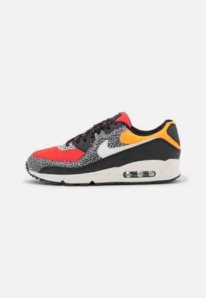 La zapatilla Nike Air Max 90 presenta una parte superior texturizada en negro y gris, con acentos rojos y amarillos, y una unidad de amortiguación de aire visible en el talón.