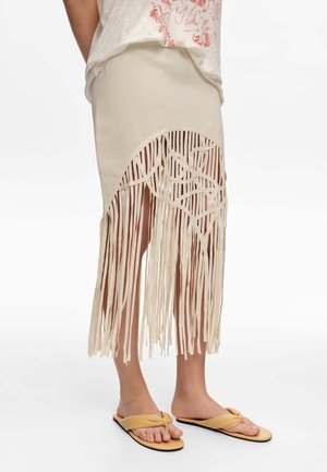 Dolna połowa osoby ubranej w beżową spódnicę z długimi frędzlami i beżowe sandały, stojąca na białym tle.