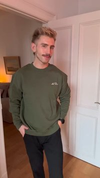 Joven con cabello castaño claro y bigote, vestido con sudadera verde y pantalones negros, de pie en una puerta con las manos en los bolsillos.