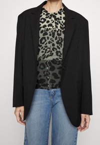 Blazer oversize nero sopra un maglione verde con stampa leopardata. Jeans blu con leggeri strappi. Il blazer presenta rever a punta e bottoni.