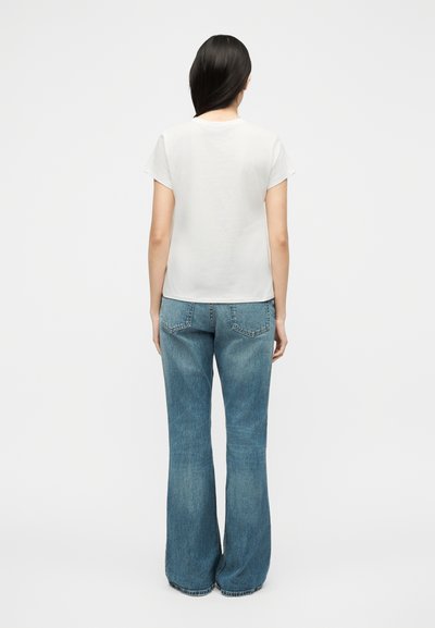 Γυναίκα με μακριά σκούρα μαλλιά που φοράει ένα απλό λευκό κοντομάνικο πουκάμισο και μπλε τζιν, στέκεται με την πλάτη προς τα εμπρός σε λευκό φόντο.