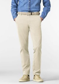 Man in beige broek met gevlochten riem, beige sneakers en een blauw denim overhemd, met handen in de zakken, staand op een witte achtergrond.