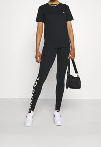 Sort bomulds t-shirt, sorte leggings med hvid "MANGOL" bogstaver, hvide sneakers, sølv kæderek, sort skuldertaske med rem.