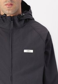 Homme portant une veste à capuche zippée gris foncé avec un petit écusson blanc rectangulaire sur la poitrine portant la mention "Element Skateboard Co.".