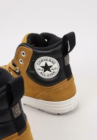 Zapatillas altas de ante marrón y cuero negro con suela de goma blanca. Presentan un parche redondo con un logo y múltiples ojales de metal para los cordones.