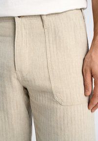 Beige, broek met visgraatpatroon gemaakt van gestructureerde stof. Voorzien van een enkel achterzak en een knoopsluiting bij de taille.