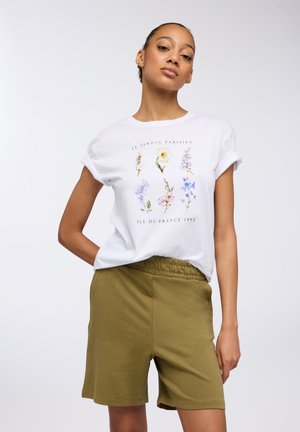 T-shirt bianca con un design floreale che include fiori viola e gialli, abbinata a pantaloni corti verde oliva con vita elasticizzata.