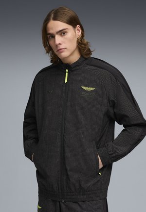 Junger Mann mit langen Haaren, der eine schwarze, gemusterte Jacke mit Reißverschluss und gelbem Logo trägt, die Hände in den Taschen, vor grauem Hintergrund.