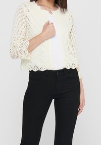 Knit cardigan in cream with openwork pattern, cropped design, scalloped hem, and three-quarter sleeves, over a white top and black jeans.