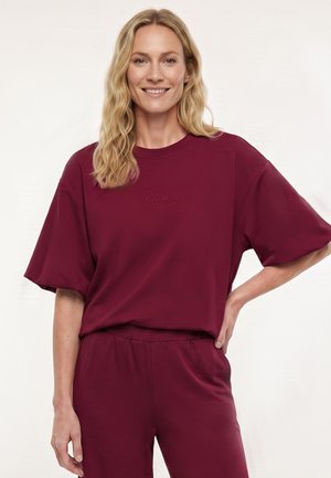 Blonde vrouw die glimlacht, draagt een losse bordeauxrode top met korte mouwen en bijpassende broek, staand met de hand op de heup tegen een lichte achtergrond.
