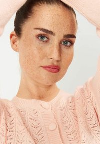 Vaaleanpunainen neulecardigan, jossa on pitsikuosi, pyöreä kaula-aukko ja napitus edessä. Pehmeä rakenne ja hienovaraiset yksityiskohdat koko vaatteessa.