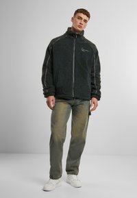 Fleece jacket in dark gray with high collar, full front zipper, and white stitching accents. Paired with faded green jeans and white sneakers.