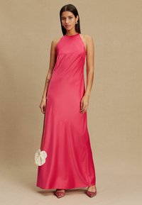 Rózsaszín szatén halter nyakú maxiruha sima sziluettel, minimális gyűrődéssel és oldalsó sliccel, rózsaszín díszített magassarkúval párosítva.
