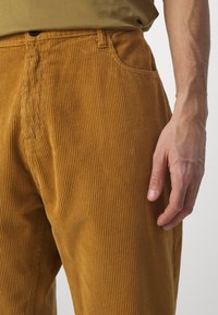 Pantalones de pana marrón con rayas verticales, que cuentan con un bolsillo delantero y una cinturilla visible con un botón. Mano descansando sobre el muslo.