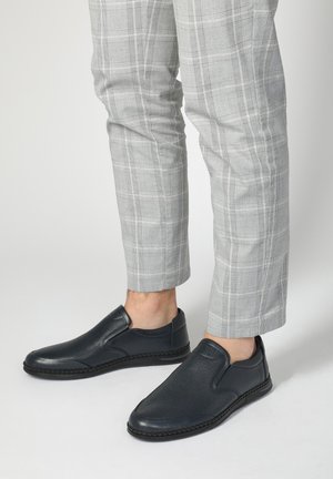 Zwarte instappers van gestructureerd leer, met een gladde afwerking en contrasterende stiksels, gedragen met grijs geruite broek.