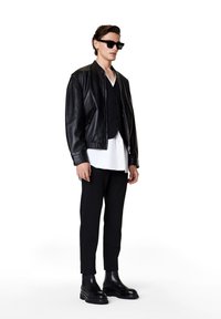 Chaqueta bomber de cuero negra sobre un chaleco negro, combinada con una camisa blanca y unos pantalones negros ajustados. Botas negras gruesas y gafas de sol rectangulares.