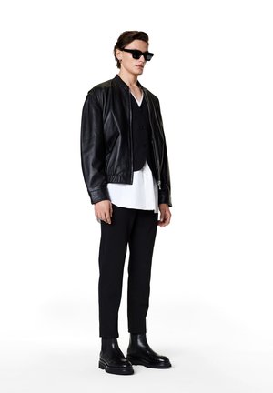 Black leather bomber jacket over black vest, paired with white shirt and slim black trousers. Black chunky boots and rectangular sunglasses.