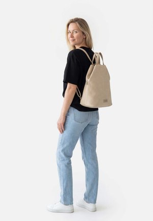 Kvinde iført sort skjorte og lyseblå jeans står og ser tilbage, bærende på en beige læderrygsæk og hvide sneakers.