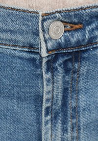 Tecido de ganga azul-claro com fecho visível de botão e costura contrastante em laranja. A cintura apresenta uma textura desfiada.