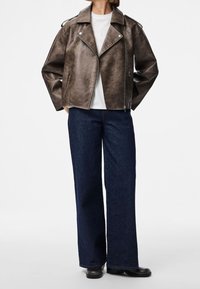 Braune Kunstleder-Bikerjacke mit breiten Revers und Reißverschlusstaschen, kombiniert mit lässigen dunkelblauen Jeans und schwarzen Stiefeln.