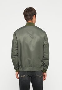 Giacca bomber verde con una superficie liscia, polsini e orlo elastici, caratterizzata da un colletto a coste e una tasca laterale sulla manica sinistra.