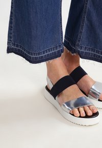 Des sandales noires et argentées avec de larges brides, un dessus argenté métallisé, un soutien élastique noir et une épaisse semelle en caoutchouc blanche ; design décontracté et moderne.