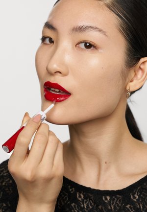 Red lip gloss is applied with a doe-foot applicator. The model has smooth skin and wears a black lace top with subtle patterns.