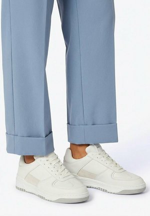 Witte lage sneakers met veters gedragen met lichtblauwe omgeslagen broek op een neutrale achtergrond.