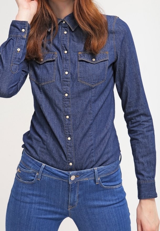 Kvinna klädd i en mörkblå denimskjorta med knappar och två bröstfickor, i kombination med medelblå denimjeans, mot en neutral bakgrund.