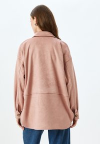 Långärmad, oversized jacka i mjukt, rosa tyg. Har en krage, knäppte manschetter och en rundad nederkant. Slät textur med minimal design.