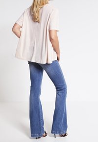 Kvinna i ljusrosa kortärmad topp, blå vida jeans och svarta klacksandaler som står mot en enkel vit bakgrund.