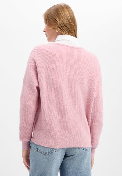 Femme aux cheveux châtain clair portant un pull rose doux et un jean bleu clair, vue de dos sur un fond blanc uni.