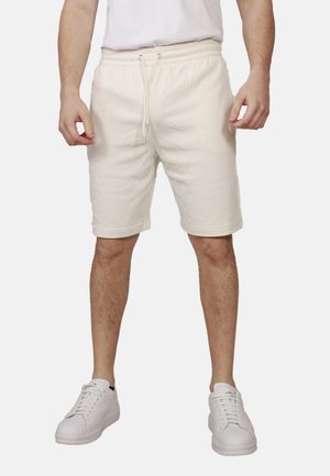 Uomo che indossa pantaloncini con coulisse color avorio, sneakers bianche e t-shirt bianca, in piedi contro uno sfondo chiaro.