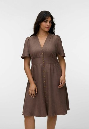 Femme aux longs cheveux foncés portant une robe marron longueur genoux à manches courtes avec une rangée de boutons sur le devant, sur un fond uni.