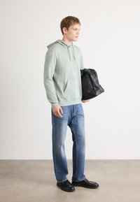 Jonge man in een lichtgroene hoodie, blauwe jeans, zwarte schoenen, met een zwarte rugzak, staand op een effen achtergrond en kijkend naar rechts.
