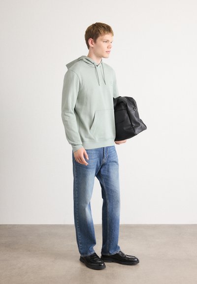 Jeune homme portant un sweat à capuche vert clair, un jean bleu, des chaussures noires, tenant un sac à dos noir, debout sur un fond uni, tourné vers la droite.
