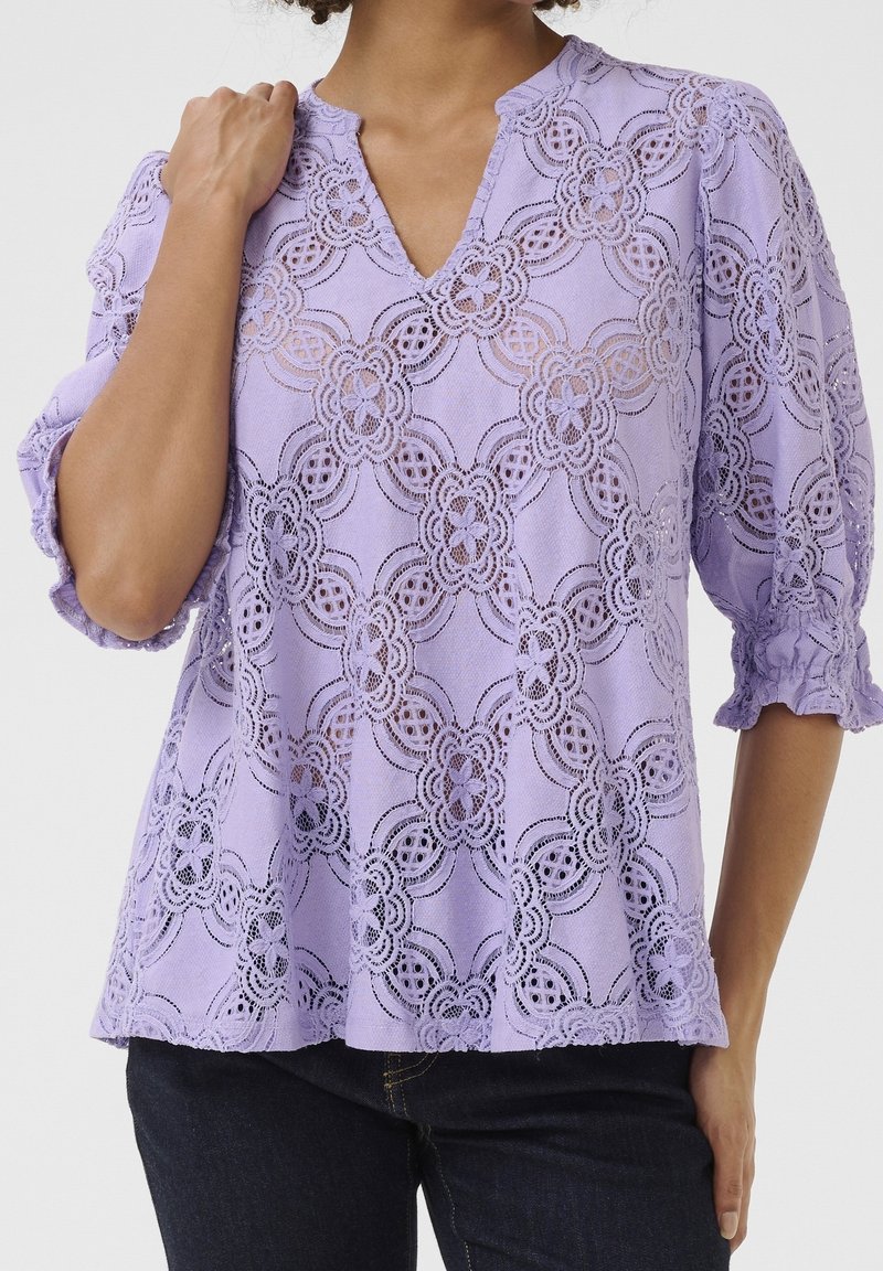 Kobieta w lawendowej koronkowej bluzce w kwiatowe wzory z trójczwartkowymi rękawami zakończonymi falbanami, w połączeniu z ciemnymi dżinsami.