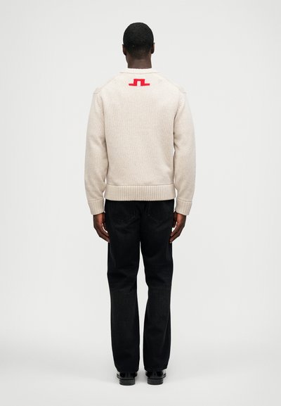 Vyras, vilkintis smėlio spalvos megztinį su raudonu logotipu ant viršutinės nugaros dalies, juodais džinsais ir juodais batais, stovi atsukęs nugarą prieš paprastą foną.