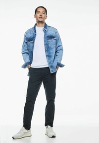 Chaqueta de mezclilla con dos bolsillos en el pecho, color azul claro, llevada sobre una camiseta blanca y pantalones oscuros. Unas deportivas blancas deportivas completan el look.