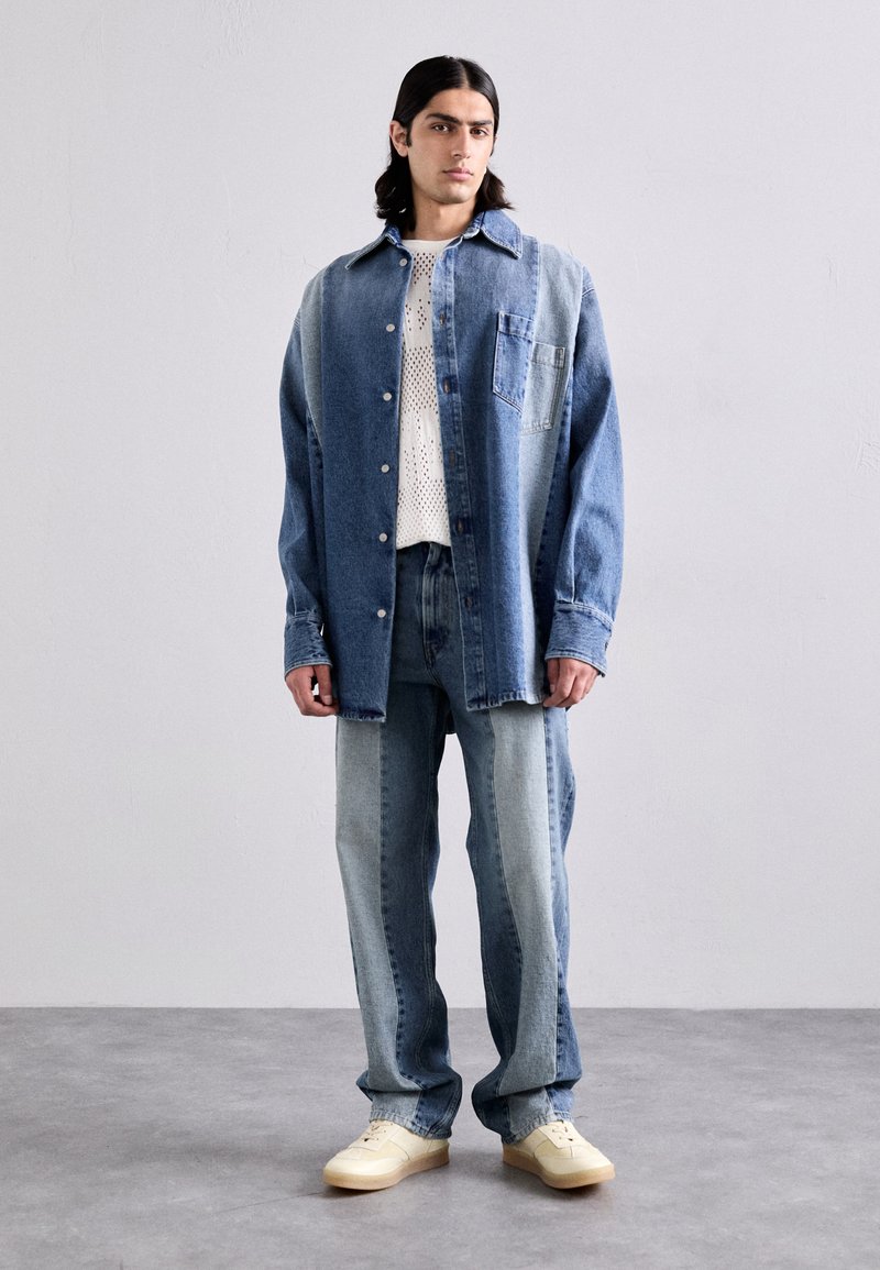 Camicia di denim, con blocchi di colore blu chiaro e scuro, design con bottoni e taschino sul petto, abbinata a jeans blu chiaro e sneakers color crema.