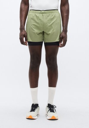 Lichtgroene korte broek met een zwarte tailleband en binnenvoering. Draagt witte sokken tot aan de kuit en sportschoenen met zwarte en oranje accenten.