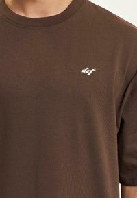 Brun bomull t-skjorte med rund hals og korte ermer, med en liten hvit logo tekst på venstre side av brystet.