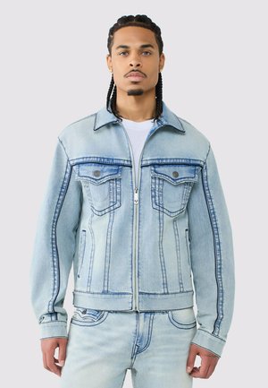 Junger Mann mit geflochtenem Haar, der eine hellblaue Jeansjacke und passende Jeans trägt, steht vor einem schlichten hellgrauen Hintergrund.