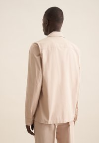 Camicia beige con bottoni, in morbido cotone. Colletto classico, maniche lunghe con polsini a bottone, design semplice e vestibilità comoda.