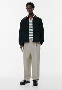 Schwarze Jacke mit braunem Kragen, gestreiftes Poloshirt in Weiß, Grün und Pink, beiger weiter Hose und schwarze klobige Schuhe.