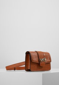 Ruskea tekokrokotiilinnahkainen ristiselkäreppu, jossa on magneettinen läppä, kultasävyinen leijonapehmeys ja säädettävä hihna.