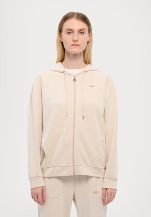 Haut à capuche beige zippé avec une texture lisse, des cordons de serrage et un petit logo. Présente des poches kangourou et des côtes aux poignets.