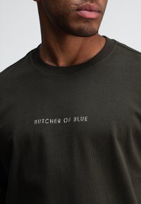 Dunkelgrünes Baumwoll-T-Shirt mit Rundhalsausschnitt und kurzen Ärmeln, auf dem "BUTCHER OF BLUE" in einer helleren Schriftart über die Brust gedruckt ist.