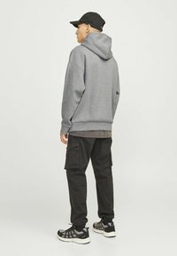 Mies harmaassa hupussa, mustissa kuorihousuissa, mustassa lippalakissa ja mustavalkoisissa tennareissa seisoo sivuttain yksinkertaisen vaalean taustan edessä.